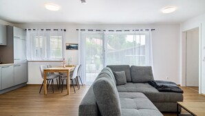 Una sala de estar moderna con un sofá seccional gris, una mesa de comedor de madera con sillas, y una zona de cocina con armarios blancos y un ventanal con cortinas traslúcidas.