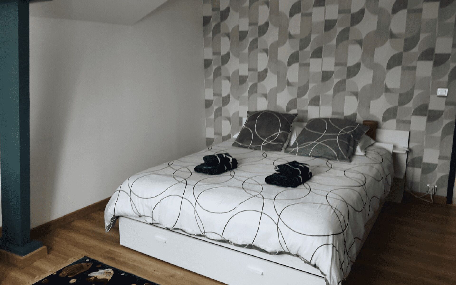 A bedroom with a patterned wall, a bed with a white headboard, and two pillows.