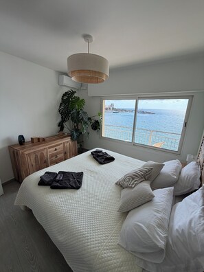 Un dormitorio con una cama grande, un aparador de madera, una planta en maceta y vista al mar a través de una ventana.