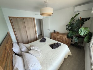 A bedroom with a bed, pillows, a wooden headboard, a wardrobe, a bedside table, a hanging plant, and an air conditioning unit.