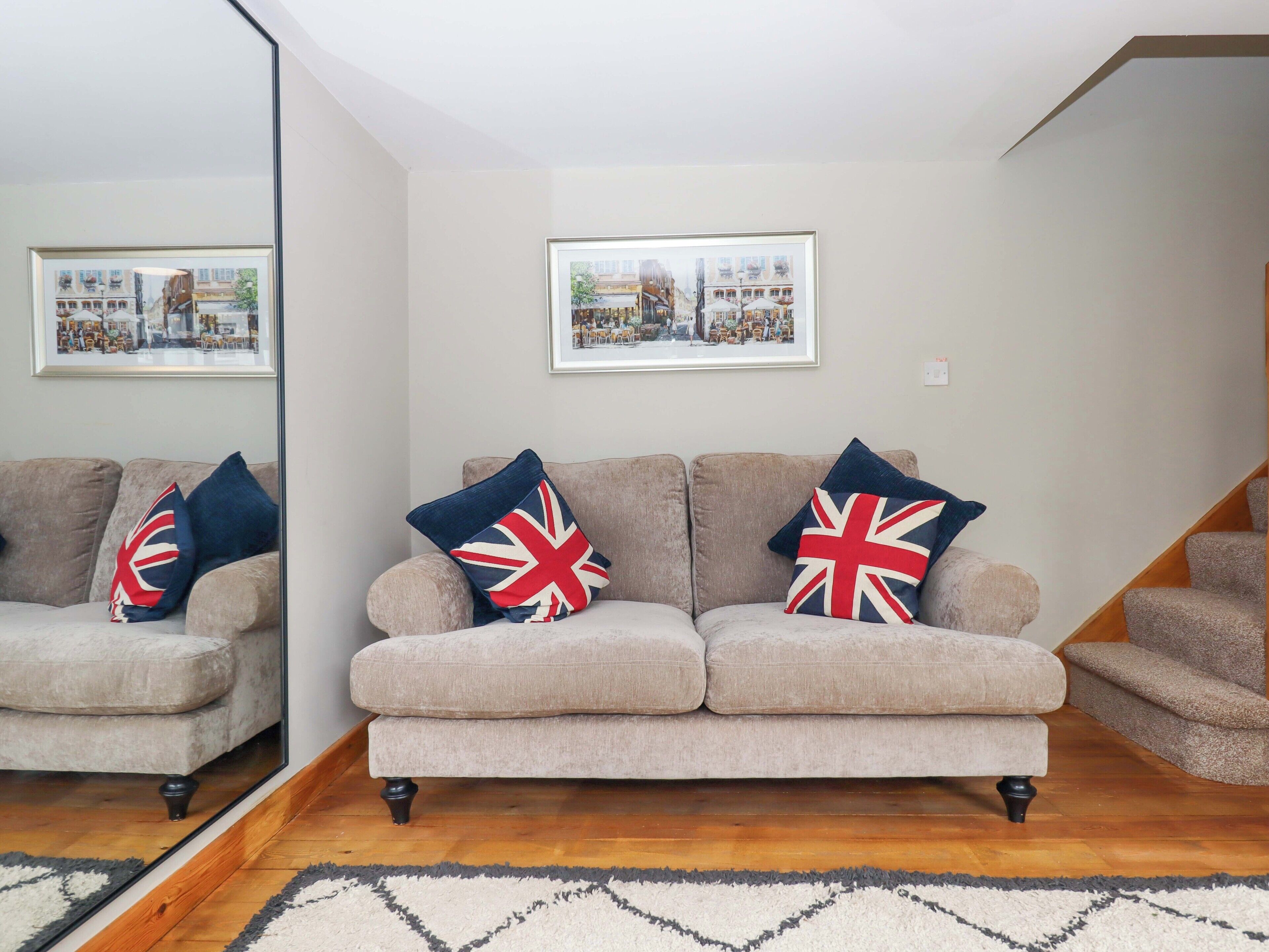 A living room with a beige sofa, two cushions with Union Jack designs, a framed cityscape picture, and a wooden staircase.