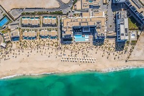 An aerial view of a resort with multiple buildings, a large sandy beach, and a swimming pool.