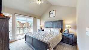 Une chambre à coucher comprenant un lit, des tables de chevet, un ventilateur de plafond, une fenêtre avec des stores et une vue sur les maisons à l’extérieur.