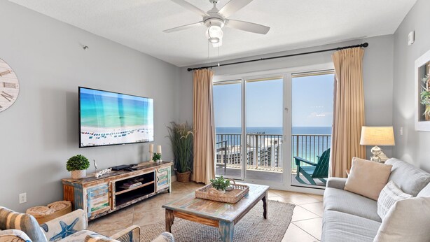 A spacious living room with a large flat-screen TV, a ceiling fan, a balcony with a view of the ocean, and a cozy seating area with a sofa and armchairs. - Breathetaking Views! New Specials! Reach out for Extended Stay Rates! (Miramar Beach)