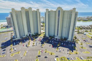 A high-rise hotel with multiple floors, a parking lot with several cars, and a view of the ocean.