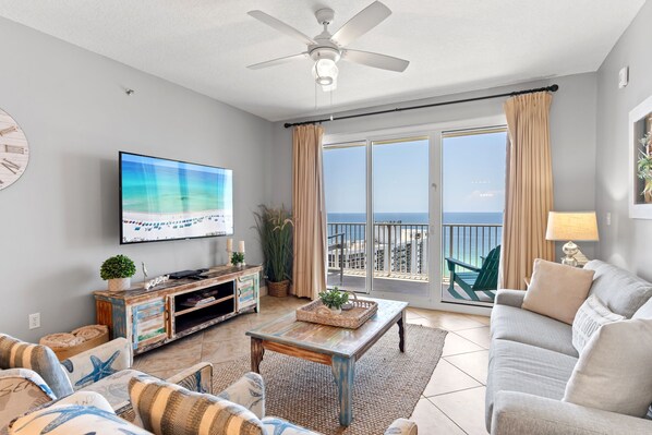 A spacious living room with a large flat-screen TV, a ceiling fan, a balcony with a view of the ocean, and a cozy seating area with a sofa and armchairs.
