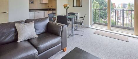 A modern living room with a leather sofa, a dining table with chairs, a kitchen in the background, and a balcony with a view of the city.