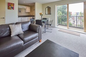 Un salon moderne avec un canapé en cuir, une table à manger avec des chaises, une cuisine en arrière-plan et un balcon offrant une vue sur la ville.