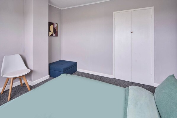 A bedroom with a bed, a chair, a small blue ottoman, a wardrobe, and a wall-mounted artwork.