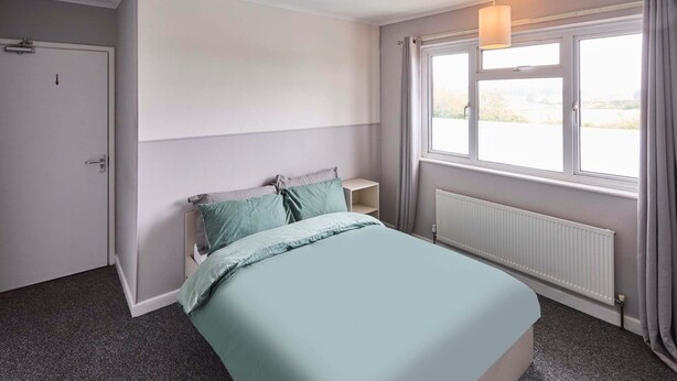Une chambre à coucher avec un lit, une table de chevet, un radiateur, une fenêtre avec des rideaux et une porte.