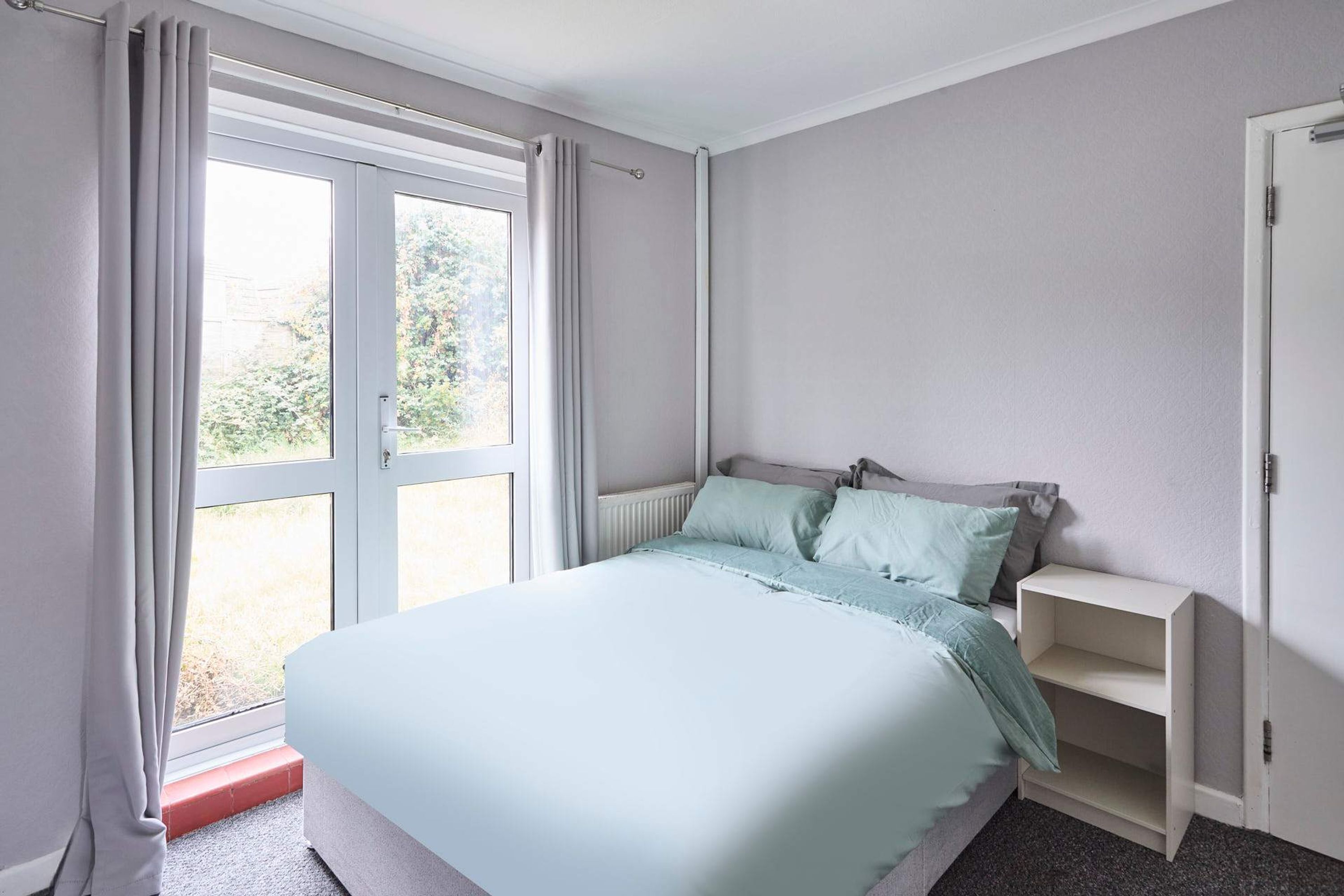 A bedroom with a bed, a radiator, a window with curtains, and a door.