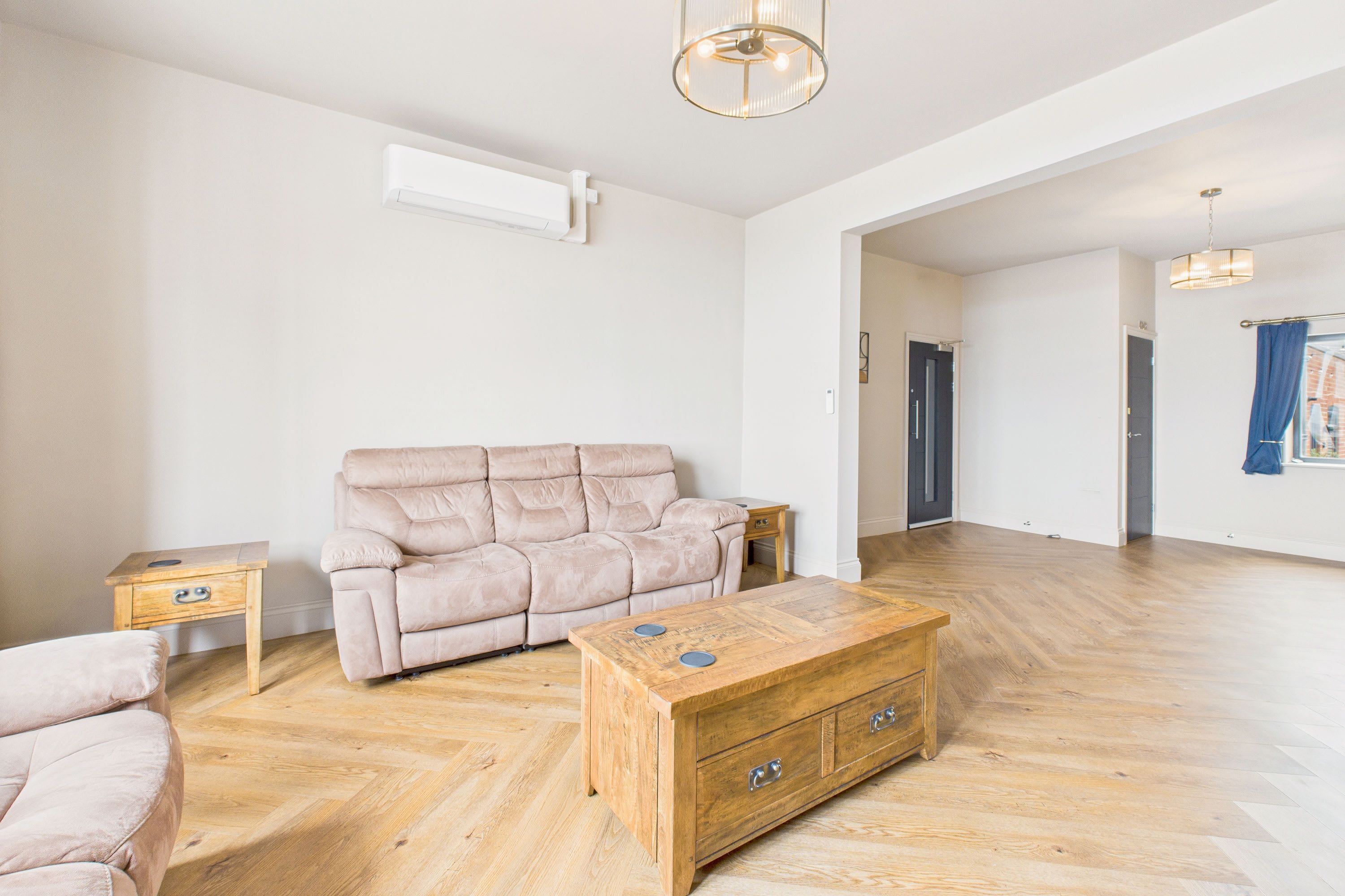 A spacious living room with a beige sofa, a wooden coffee table, and a herringbone-patterned wooden floor.