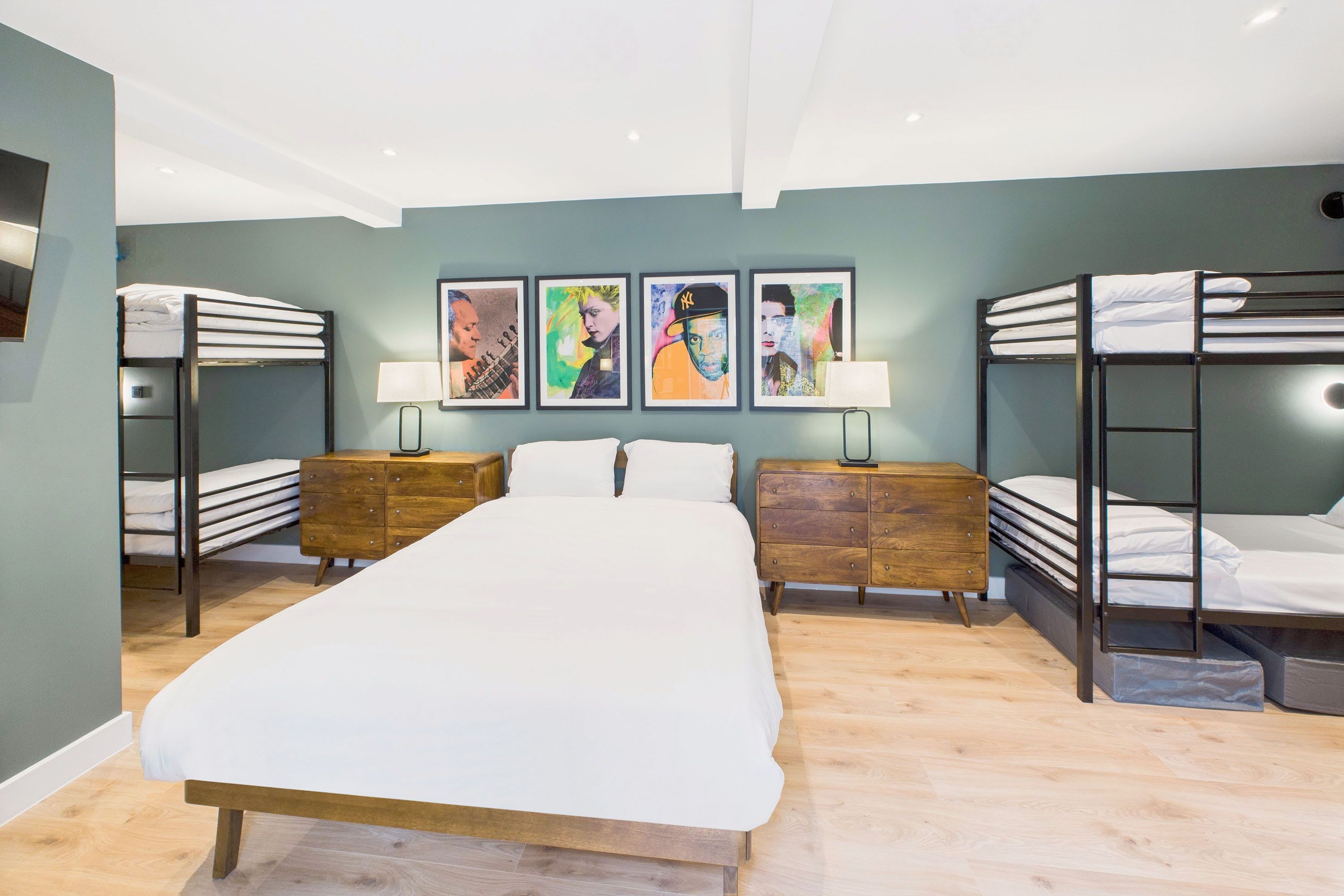 Ein modernes Hotelzimmer mit einem großen Bett, zwei Nachttischen, einer Holzkommode, Etagenbetten und vier gerahmten Kunstwerken an der Wand.