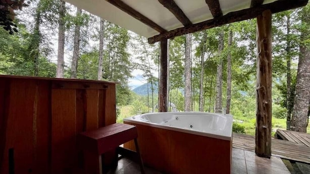Une terrasse en bois avec un jacuzzi et un banc, entourée d’arbres et offrant une vue sur la montagne.