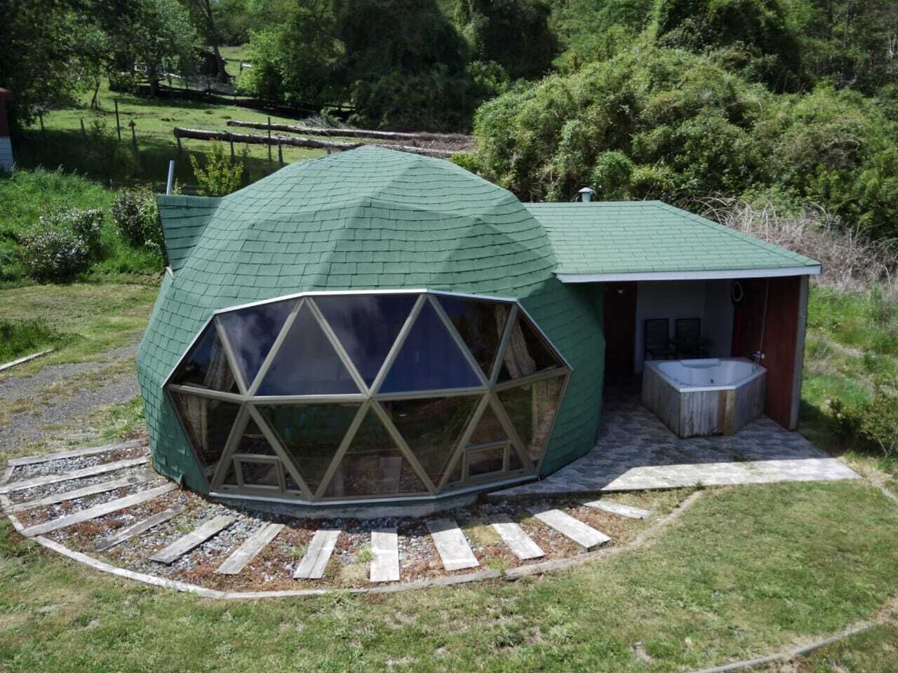 Una estructura de domo geodésico con un gran ventanal y un pequeño edificio adosado con un jacuzzi.