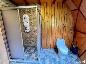A small, rustic bathroom with a stone floor, a wooden shower enclosure, and a basic toilet. - Domo Huepil (Pucon)