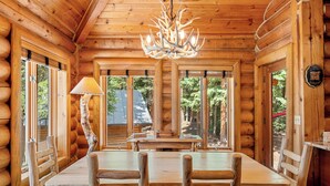A log cabin interior with a dining table, chairs, a chandelier, and large windows offering views of the surrounding forest.