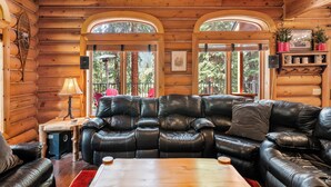 A log cabin interior with a leather sofa, wooden tables, and large windows offering views of the forest.