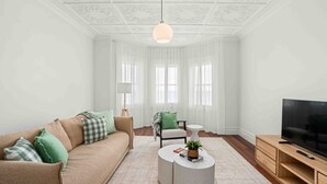 Ein modernes Wohnzimmer mit einem beigefarbenen Sofa, grün-weißen Karokissen, einem runden Couchtisch mit Pflanze, einem Fernseher auf einem Holzschrank und einem großen Fenster mit transparenten Vorhängen.