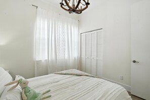 Un dormitorio con una cama, un peluche, una araña, un armario y una ventana con cortinas.