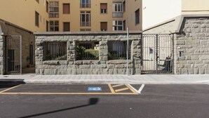 Un estacionamiento con señal de estacionamiento para discapacitados, un muro de piedra y una entrada con portón a un edificio con múltiples ventanas y balcones.
