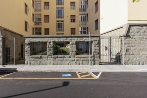 Un estacionamiento con señal de estacionamiento para discapacitados, un muro de piedra y una entrada con portón a un edificio con múltiples ventanas y balcones.
