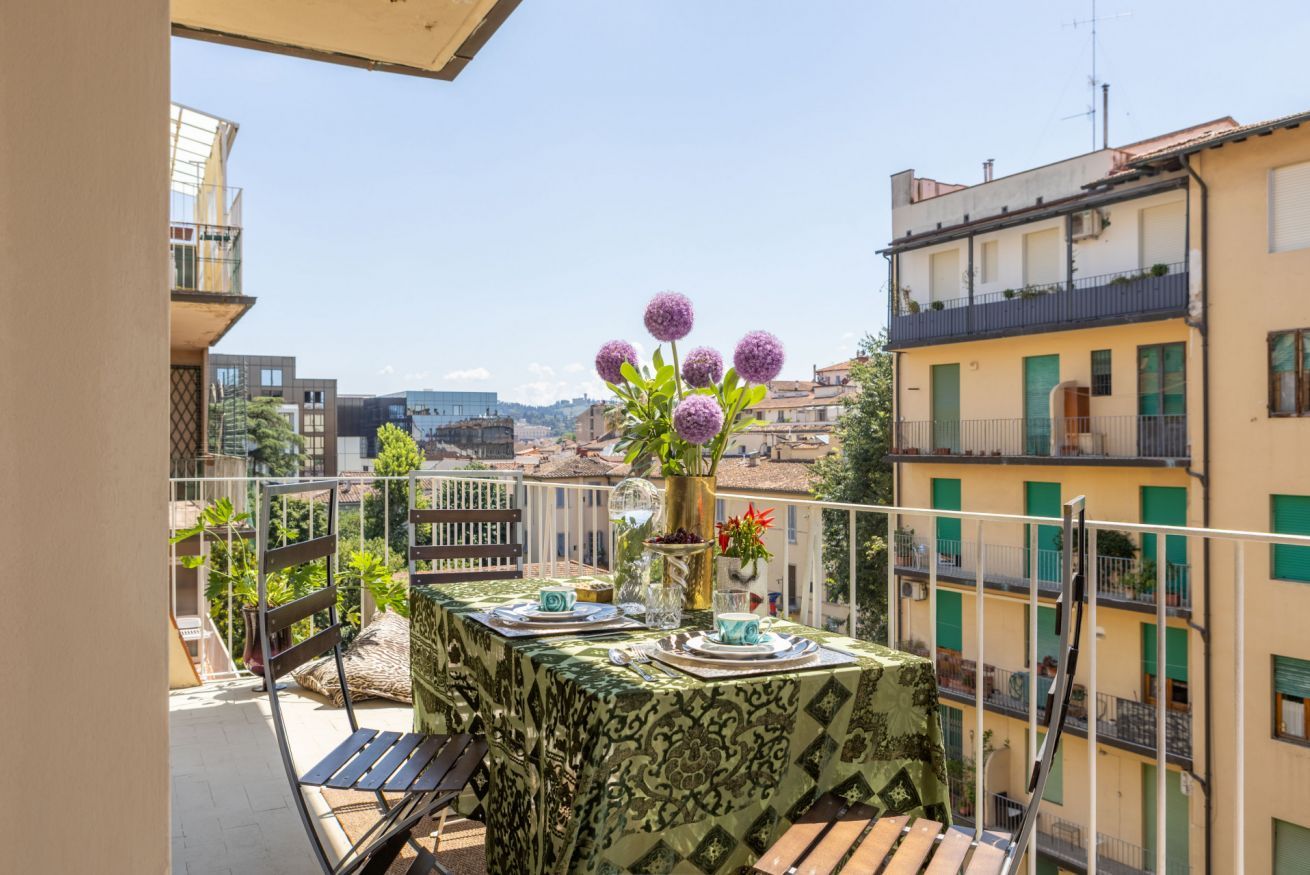 A balcony with a table set for two, a vase with flowers, and a view of the cityscape.