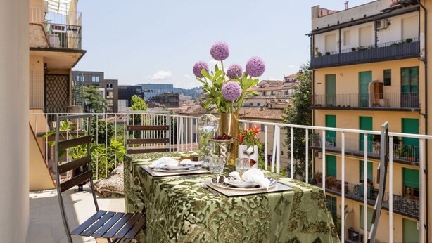 A balcony with a table set for dining, a vase with flowers, and a view of the cityscape.