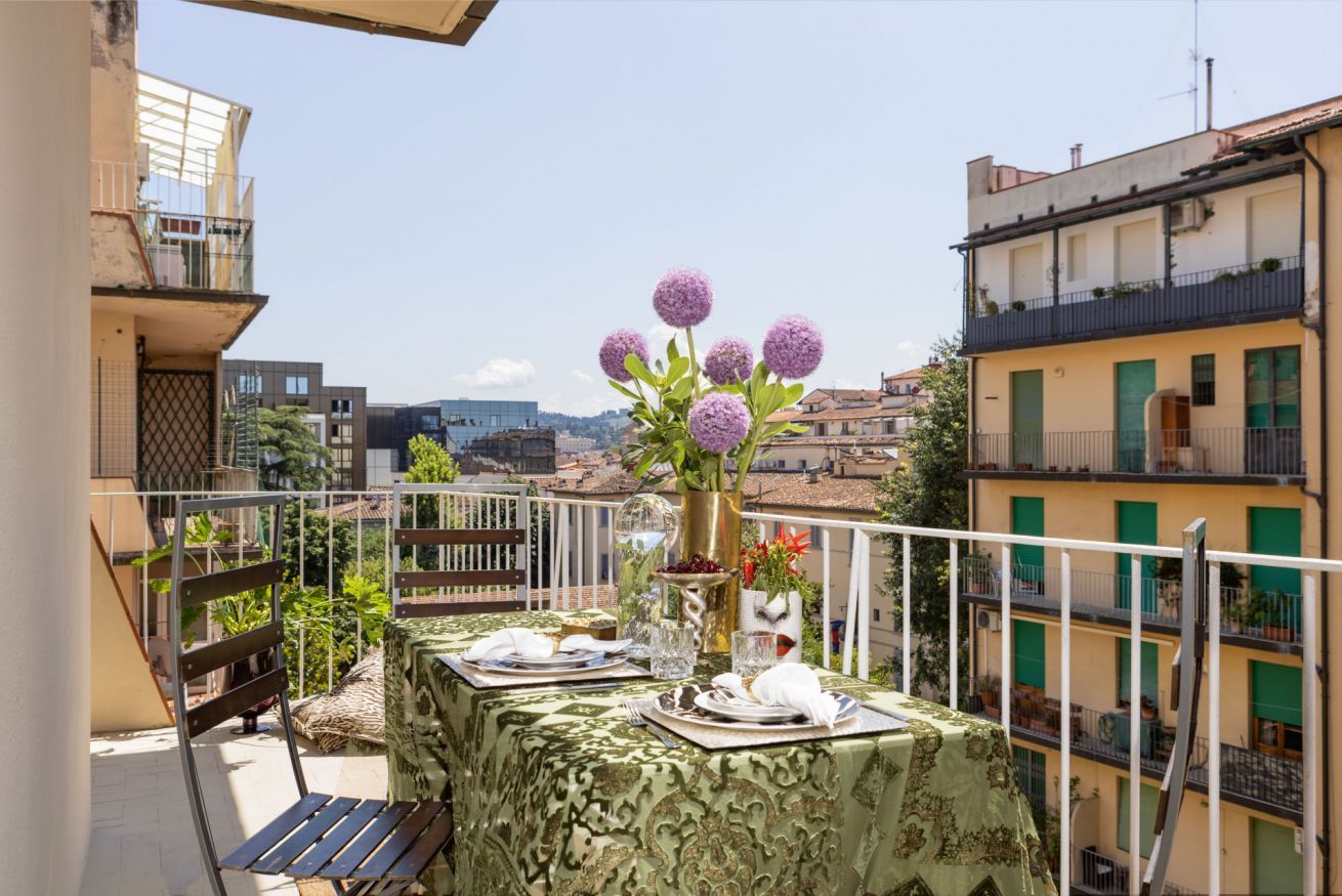 Un balcon avec une table dressée pour le dîner, un vase rempli de fleurs et une vue sur le paysage urbain.