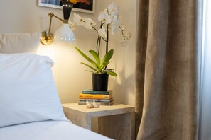 A neatly made bed with white linens, a bedside table with a potted orchid, a stack of books, and a wall-mounted lamp.