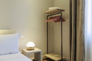 Una habitación de hotel con una cama, una mesita de noche con lámpara, un perchero y un pequeño estante con una toalla.