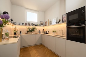 Una cocina moderna con armarios blancos, suelo de madera en espiga, horno empotrado y varios electrodomésticos.