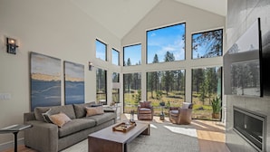A spacious living room with a large gray sofa, a coffee table, and a TV. There are multiple windows offering views of a forest and a lake. - Forestbrook Luxury with Access to Exclusive Golf (Sunriver)
