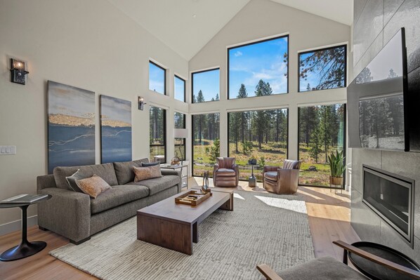 A spacious living room with a large gray sofa, a coffee table, and a TV. There are multiple windows offering views of a forest and a lake. - Forestbrook Luxury with Access to Exclusive Golf (Sunriver)