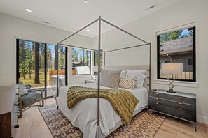 A modern bedroom with a four-poster bed, a nightstand, and a large window offering a view of trees and a cabin. - Forestbrook Luxury with Access to Exclusive Golf (Sunriver)
