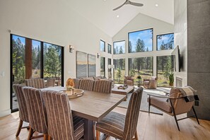 A spacious dining area with a large wooden table and striped upholstered chairs, large windows offering a view of a forested landscape, and a modern ceiling fan. - Forestbrook Luxury with Access to Exclusive Golf (Sunriver)