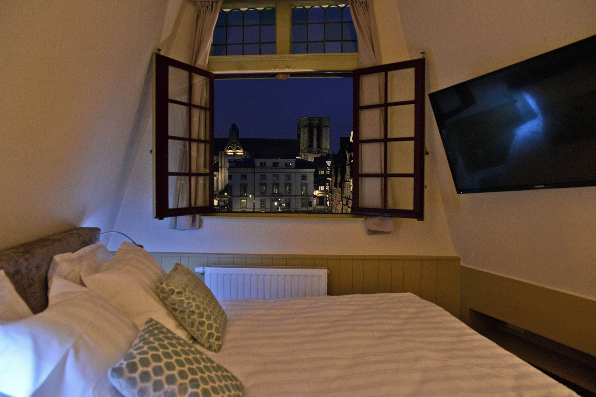 A bedroom with a large bed, a window with curtains, a television on the wall, and a view of buildings outside.