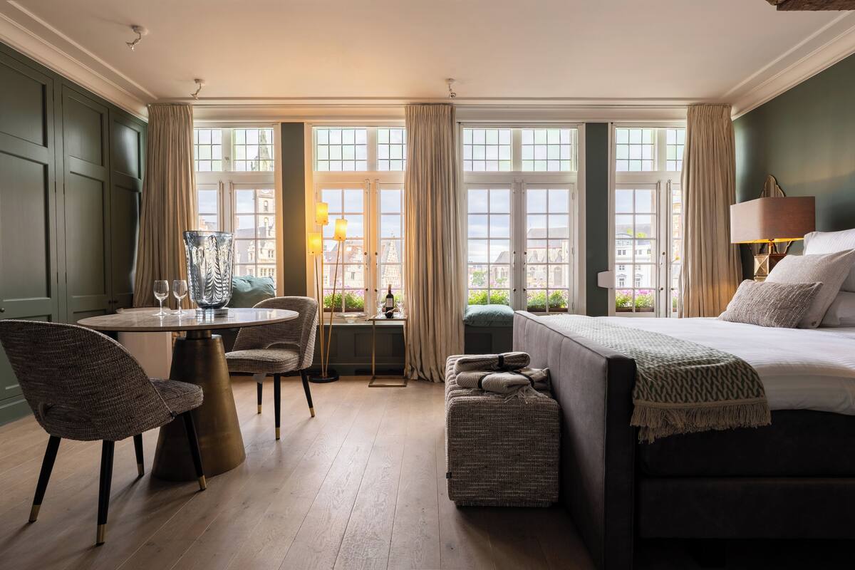 A hotel room with a bed, a sofa, a dining table with chairs, and large windows with curtains.