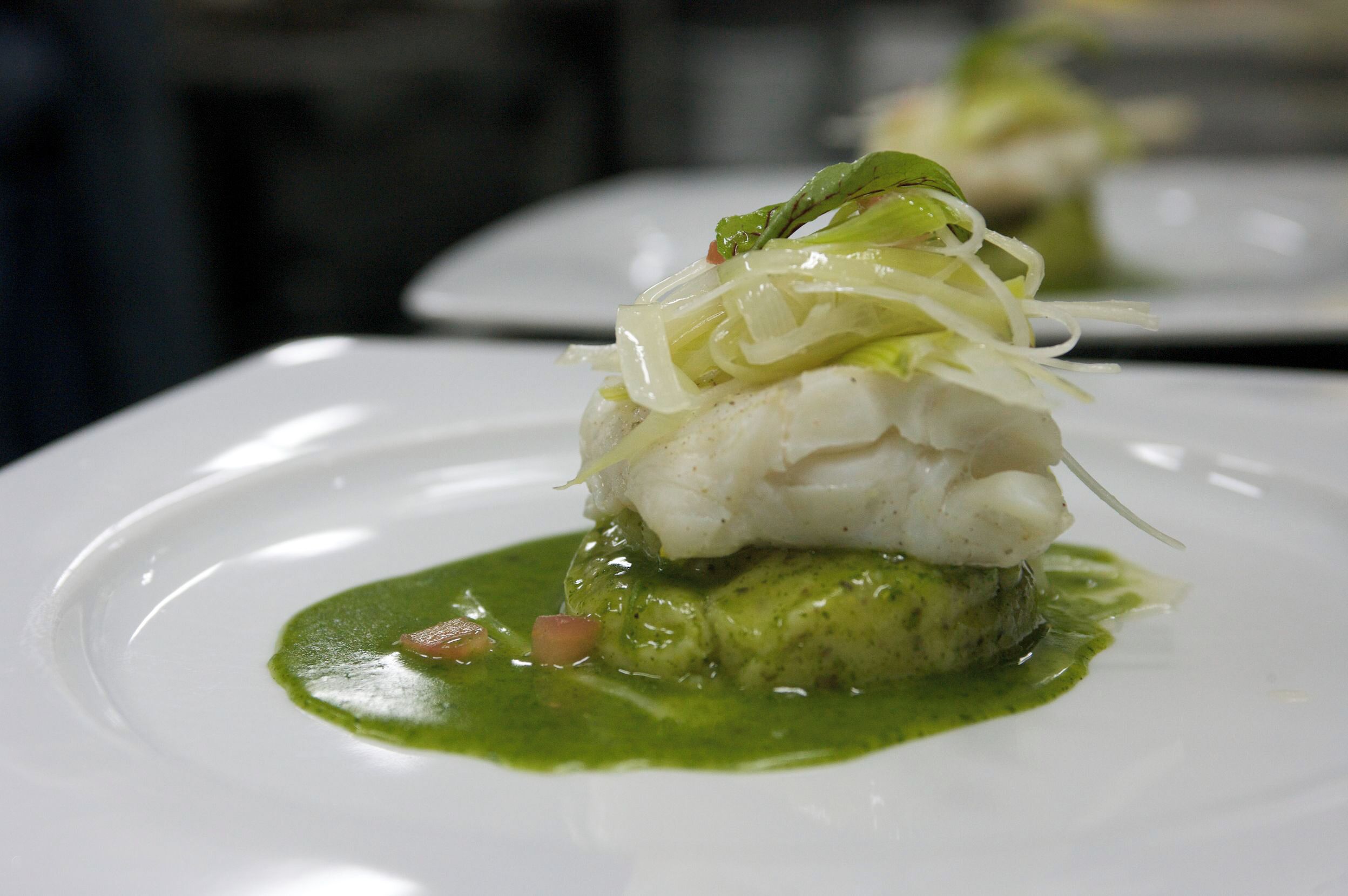 A white plate with a fish dish, green sauce, and vegetables.