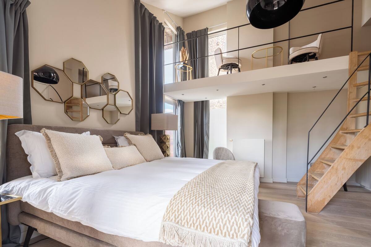 A modern bedroom with a large bed, a loft area with seating, and a staircase.