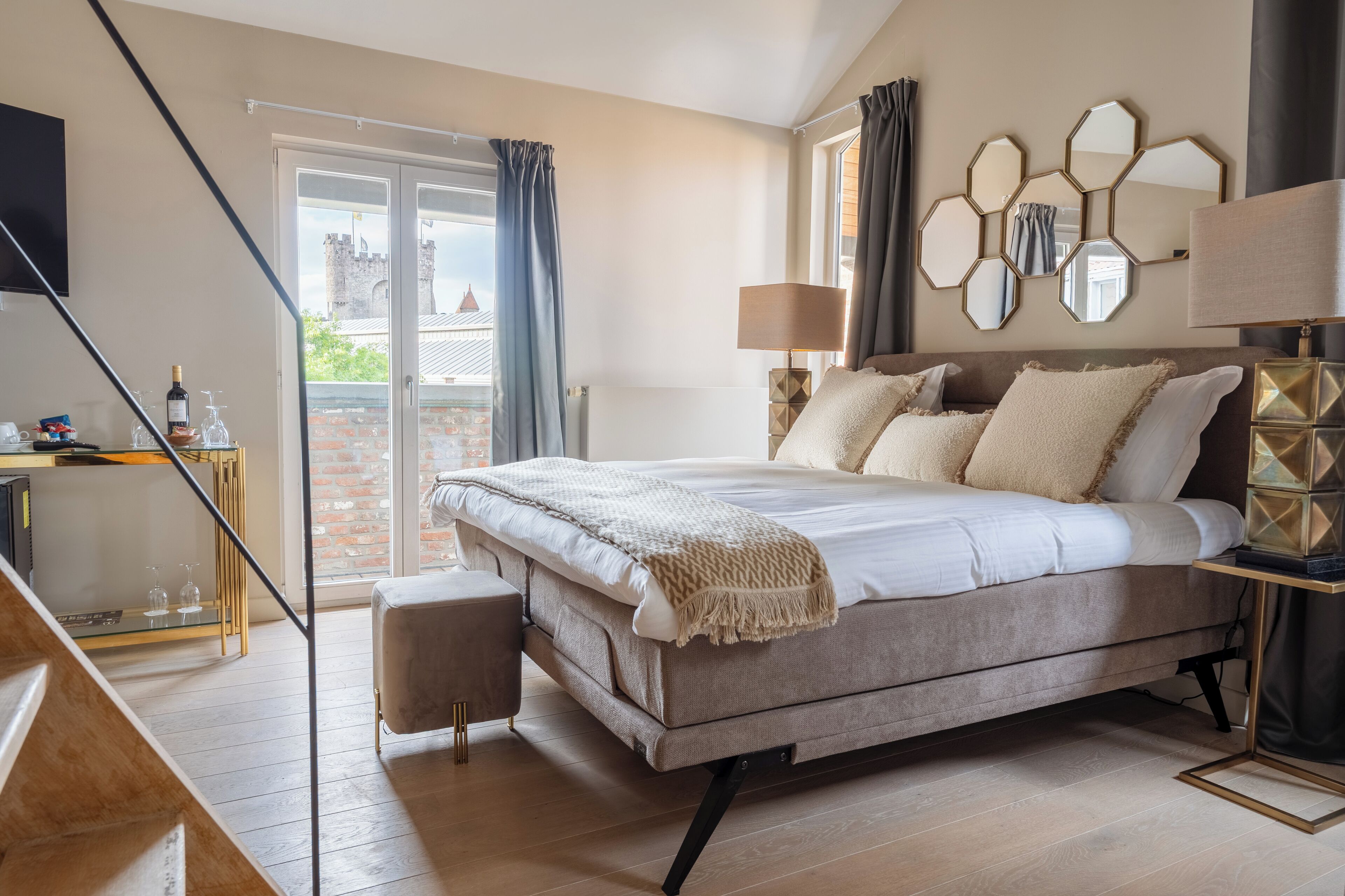 Une chambre avec un grand lit, un bureau avec une lampe, une vue sur un château par la fenêtre et un balcon avec une balustrade.