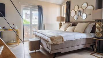Une chambre avec un grand lit, un bureau avec une lampe, une vue sur un château par la fenêtre et un balcon avec une balustrade.