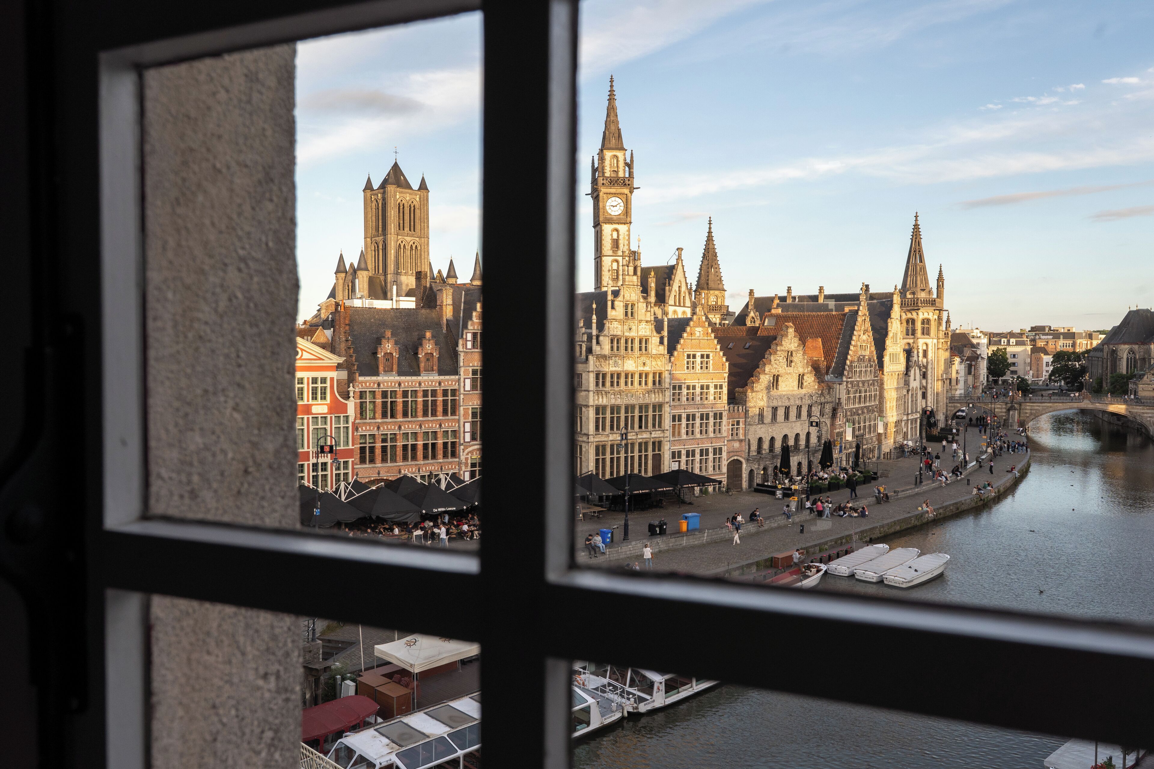 A view of a historic European cityscape through a window, featuring a prominent church with a clock tower, a river with boats, and a pedestrian walkway.