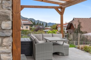 Une terrasse extérieure aménagée avec des meubles en osier, un mur en pierre et une vue sur des maisons et des montagnes en arrière-plan.