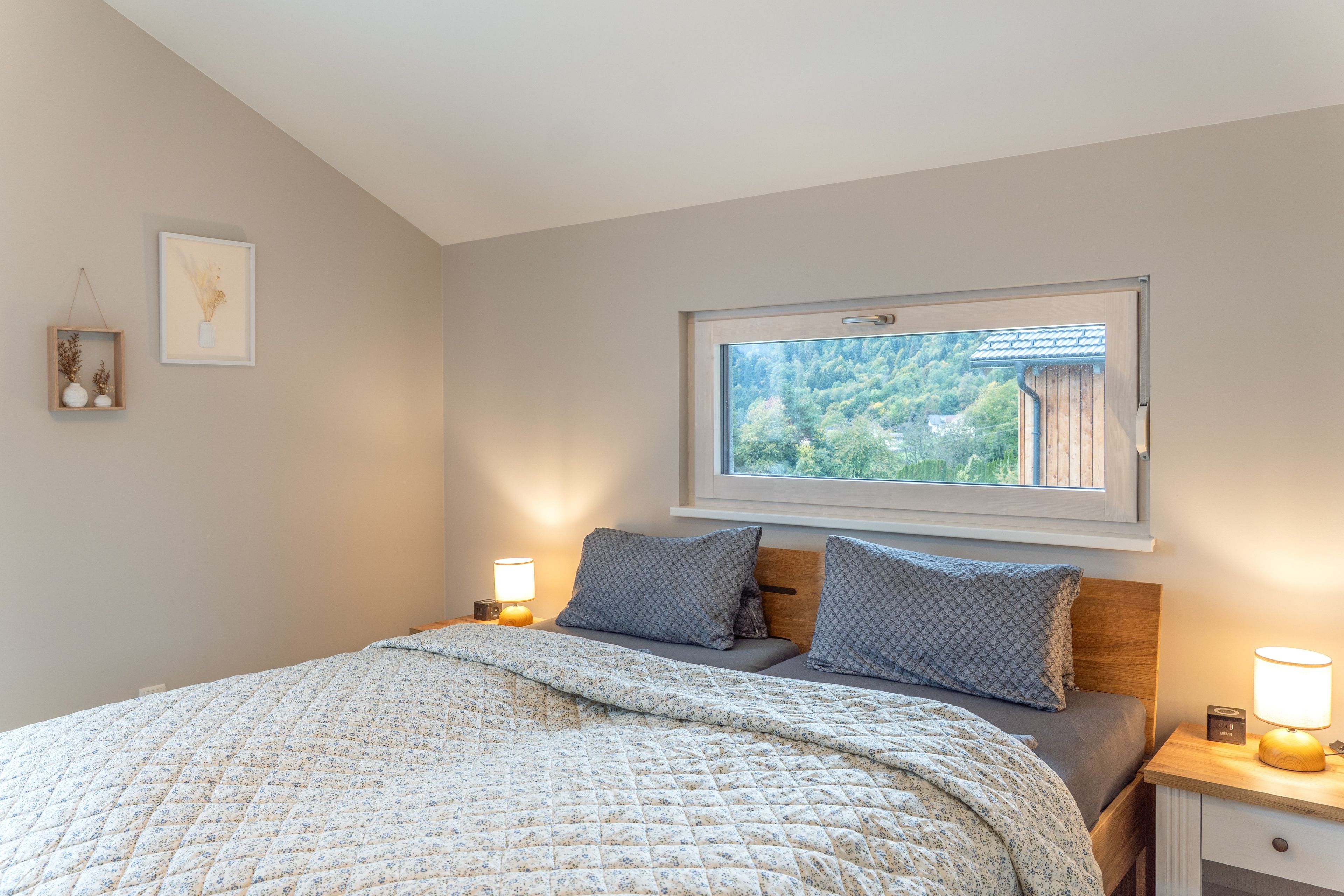 Ein Schlafzimmer mit einem Bett, zwei Nachttischen, einem Fenster mit Blick auf Bäume und einem Wandbild.