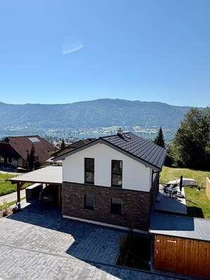 Una casa moderna con techo inclinado, un porche cubierto y vistas a un lago y montañas.