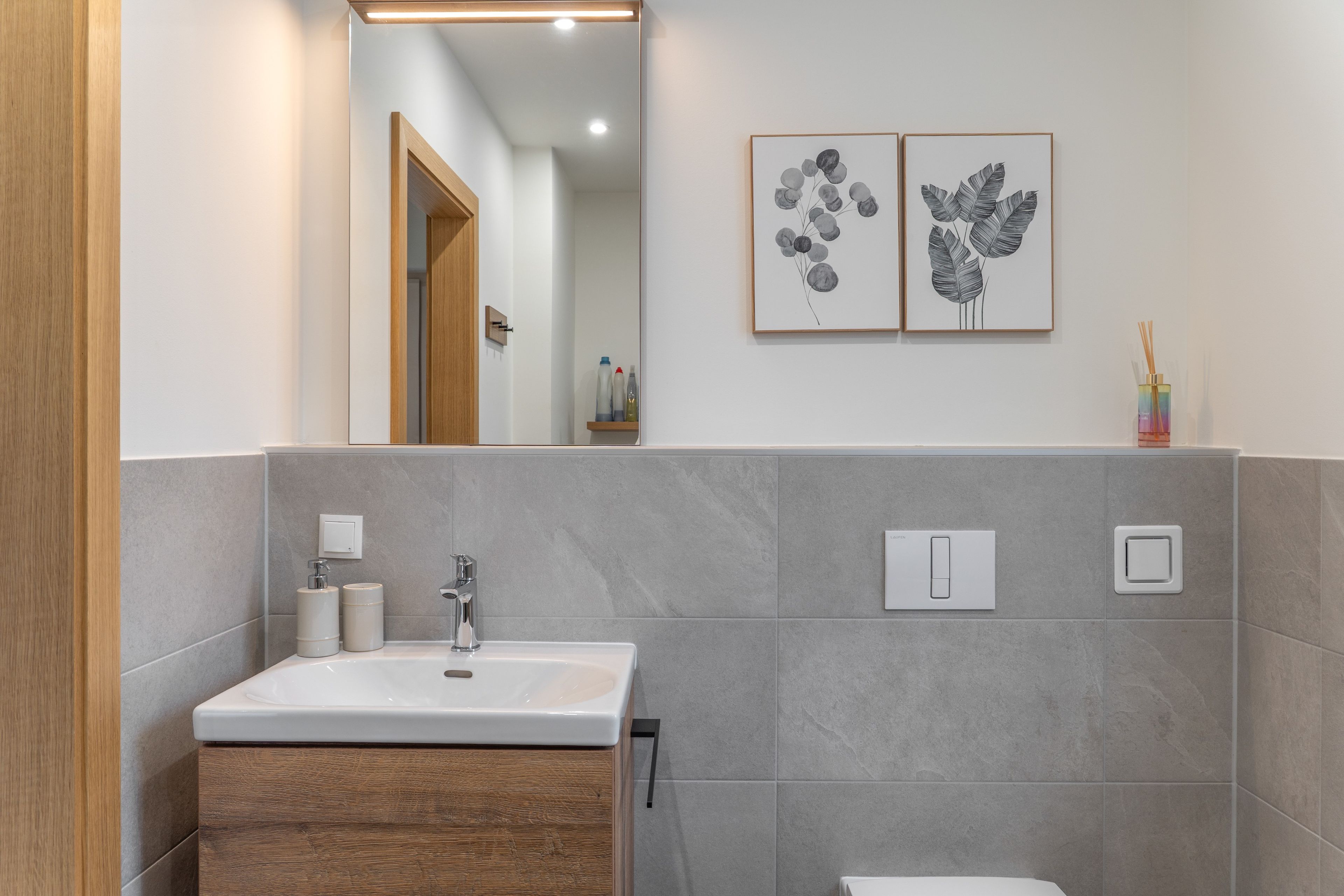 A modern bathroom with a wooden vanity, a white sink, and grey tiles. There are two wall-mounted light switches and a framed botanical print above the mirror.