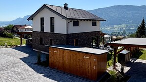 A modern two-story house with a stone facade, a wooden deck, and a view of a lake and mountains. - Holiday Home 'Steinrose' with Lake View, Private Terrace and Wi-Fi (Bodensdorf)