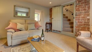 A living room with a sofa, a coffee table with a book and drinks, a brick wall, and a staircase. - Forget Me Not Cottage (Binham)
