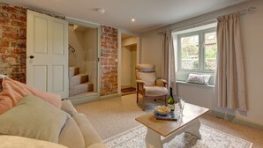 A cozy living room with a brick wall, a white door, a beige armchair, a wooden coffee table, and a window with curtains. - Forget Me Not Cottage (Binham)